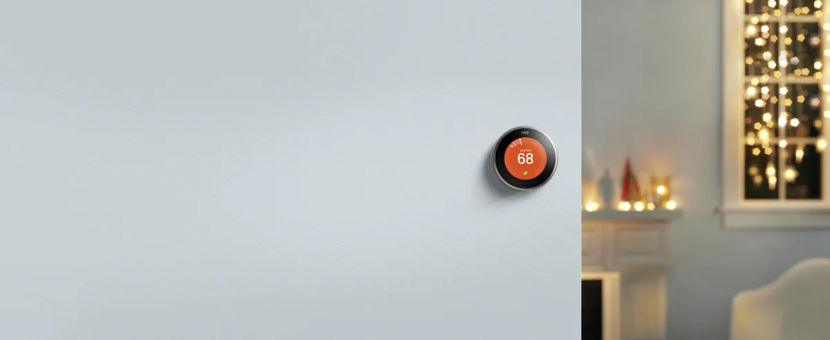 A Nest thermostat that reads “Heating — 68” hangs on a light blue wall. In the background is a fireplace and a window with dangling lights.