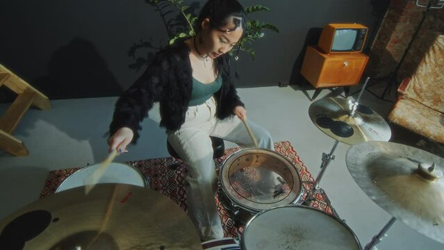 Inspired Asian female musician playing fast tempo on drum set while practicing drumming in studio. View from above