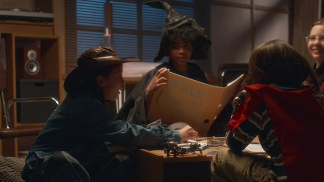 Medium shot of multiracial kids wearing costumes of wizard, magician and cowgirl playing board game at home in evening