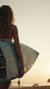 Sexy lady surfer standing on beach in sunset or sunrise, back view, vertical video. Slender lady in bikini holding surfboard and playing with her long hair, carefree woman on ocean shore, active rest