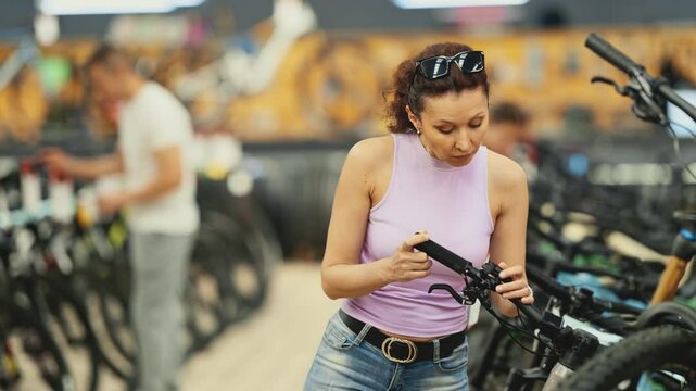 Female shopper in casual tank top and jeans carefully inspecting mountain bicycle handlebars and controls in store, showing curiosity and determination in choosing right bike for adventures