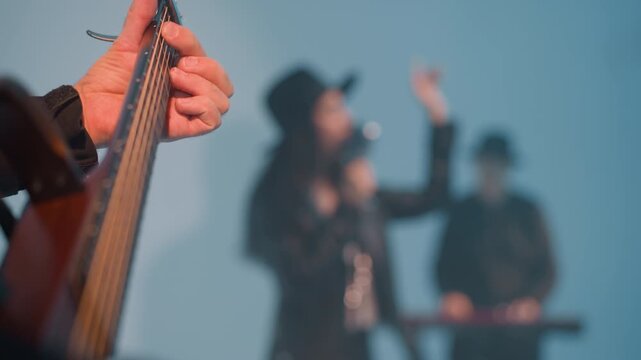 Close view of a faceless guitarist playing a guitar against a white background with a blurred singer and keyboardist in the background. The guitarist's hand is focused as they strum the strings