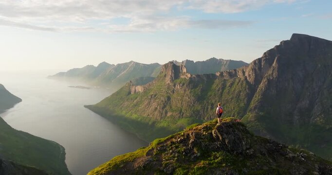 Female on arctic Norway mountain top look at fjord in sunset glow; drone reveal