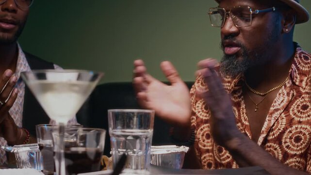 Black woman and three African American men composing new song while sitting at bar table with food and drinks talking, clapping to rhythm and making notes