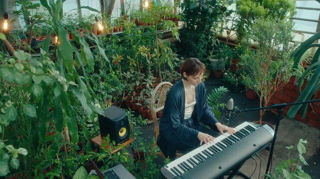 Female music artist surrounded by lush plants playing keyboard beside microphone in cozy greenhouse decorated with string lights. High angle view