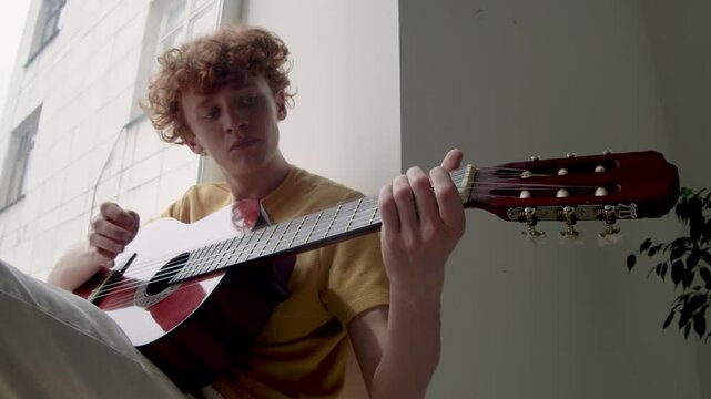 Low angle shot of gen Z Caucasian guy sitting on window sill and practicing playing acoustic guitar, learning song by himself at home