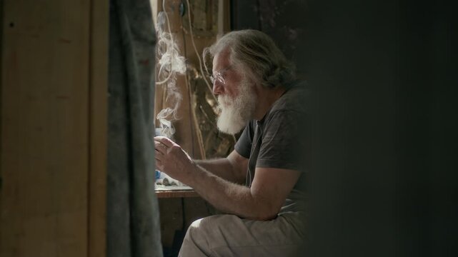 Medium shot of Caucasian senior man soldering while sitting by window in cozy small workshop