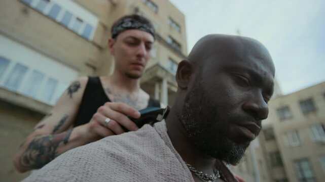 Close up view of grim-faced Black man sitting with towel on shoulders while tattooed friend shaving his head bald with electric razor on urban street