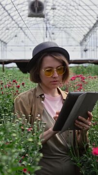 Vertical shot of female florist standing among lush plants in greenhouse, examining flowers and using app on digital tablet