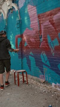 Full vertical shot of male graffiti artist in respirator creating new mural on wall of brick house in city street, adding touches of pink spray paint to red and blue background, on sunny day