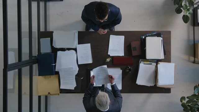 Top down view of two colleagues in office, male assistant standing in front of his boss and handing him documents, furious senior boss tearing them apart and throwing on desk