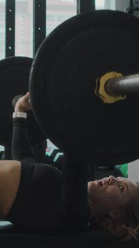 Vertical side view medium shot of athletic young Caucasian woman performing barbell bench press at gym