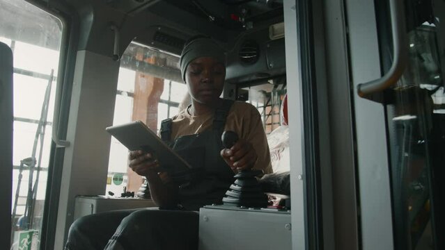 Black female quality control inspector sitting inside of tractor cabin, checking gearshift and working on digital tablet at machinery plant