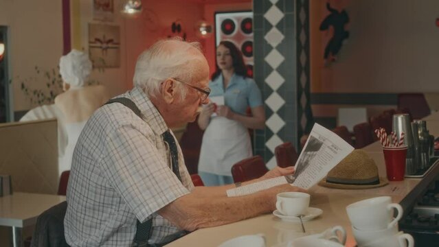 Side shot of senior Caucasian gentleman in glasses reading article in morning newspaper while sitting at counter in vintage style restaurant