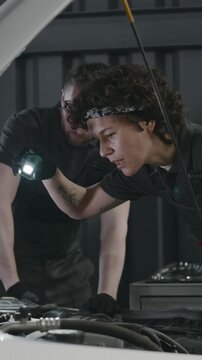 Medium vertical shot of Caucasian female mechanic in gray overalls, with torch inspecting clients car engine for damage, and male coworker with wrench removing battery for checkup