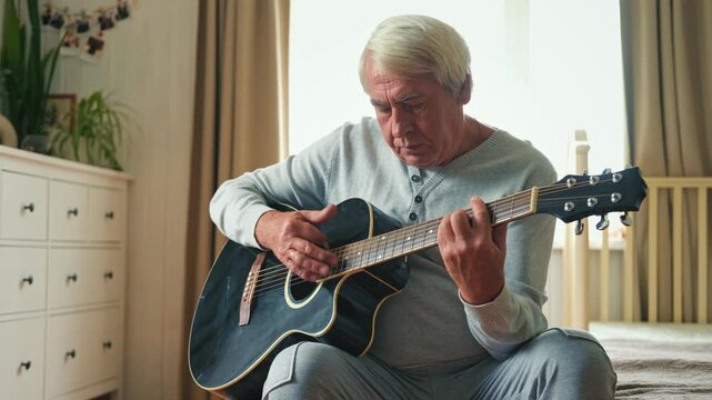 Senior man is playing guitar. Elderly man sitting on the sofa and playing guitar. Portrait of a gray-haired mature man in a sweater and glasses learning to play. Enjoying retirement life at home.