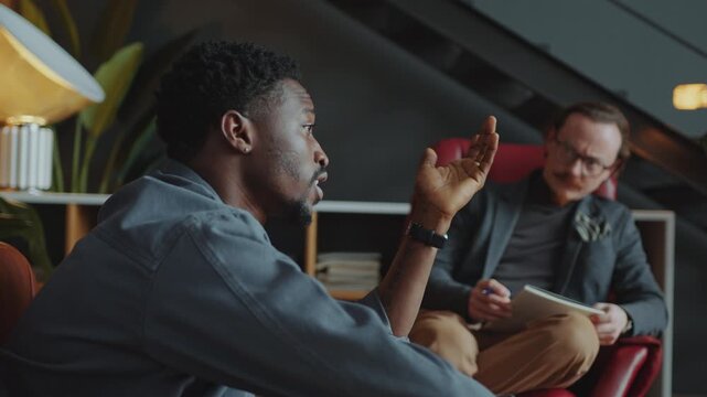 Young Black man explaining his feelings to Caucasian male therapist listening attentively, writing notes and giving advice during individual counseling session in cozy office. Rack focus