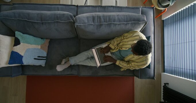 A man in casual clothes uses a laptop while sitting on the couch. Top view, unrecognizable person. Daylight