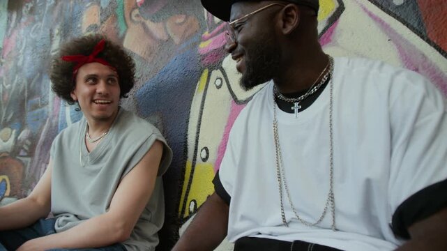 Young Black man sitting by graffiti wall outdoors in urban street, chatting with cheerful friend and giving him hand clasp