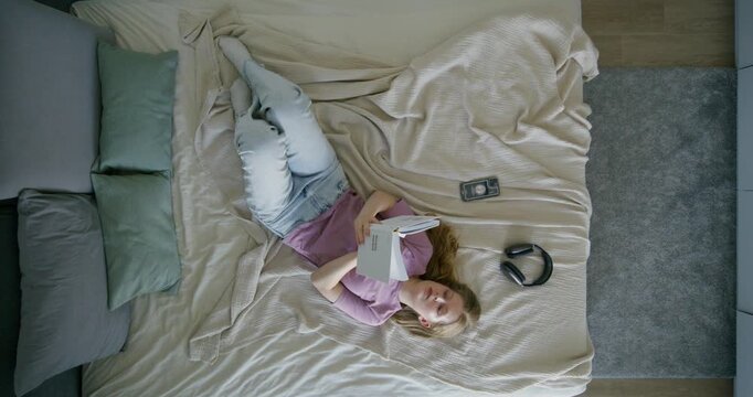 A young woman in casual clothes is reading a book lying on her bed at home. Daylight, top view