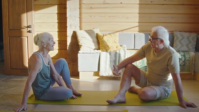 Full shot of elderly retired married couple in sports clothes sitting on mats in living room, having conversation, discussing difficulties and problems after morning exercise routine