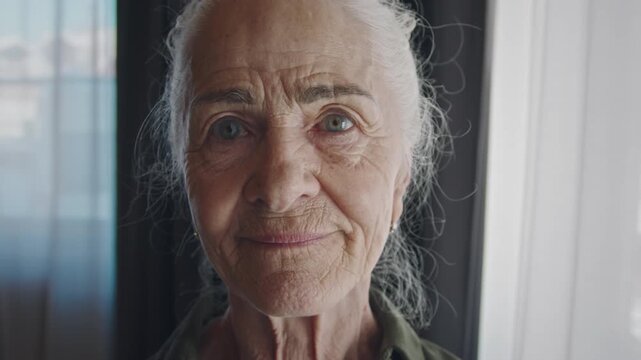 Extreme close-up face portrait of joyous elderly Caucasian woman with beautiful natural wrinkles, long grey hair, posing by window in daylight, looking at camera and smiling