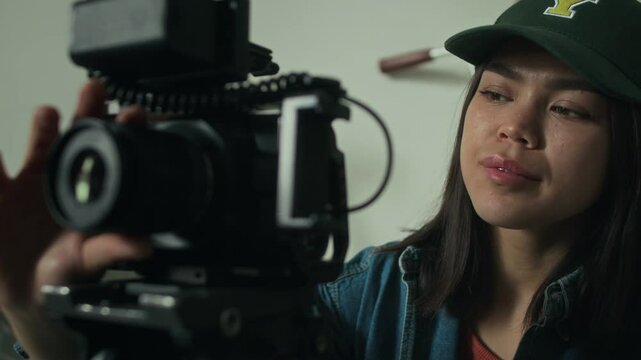 Closeup of Caucasian camerawoman in cap slightly moving camera horizontally during filmmaking process indoors