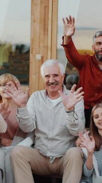 Vertical group portrait of four cheerful relatives waving at camera while sitting on terrace of wooden countryside house during outdoor family party