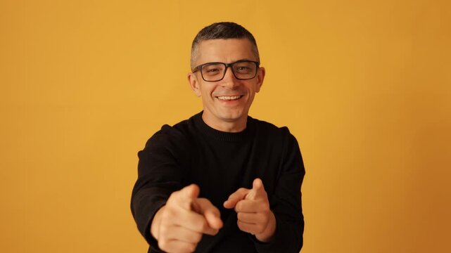 I choose you. Satisfied man in glasses wearing black jumper pointing and showing selection while looking at camera and expressing joy on isolated over yellow background