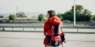 Eine Frau mit Rucksack steht mit dem Rücken zur Kamera vor oder an einer Autobahn