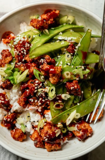 An overhead shot shows a bowl of crispy air fryer tempeh, cooked snow peas, and white rice. The food is drizzled with a gochujang glaze and garnished with sliced green onions and sesame seeds.