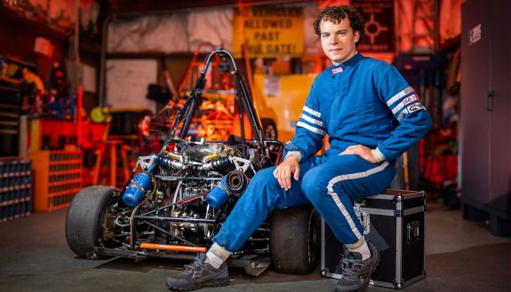 Connor Gregg in a racing jumpsuit sits next to the Motorsports club's car in a garage filled with tools