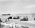 Scene on "Omaha" Beach on the afternoon of "D-Day", 6 June 1944, showing casualties on the beach, a bogged-down "Sherman" tank, several wrecked trucks and German anti-landing obstructions. A LST is beached in the left distance and invasion shipping is off shore.