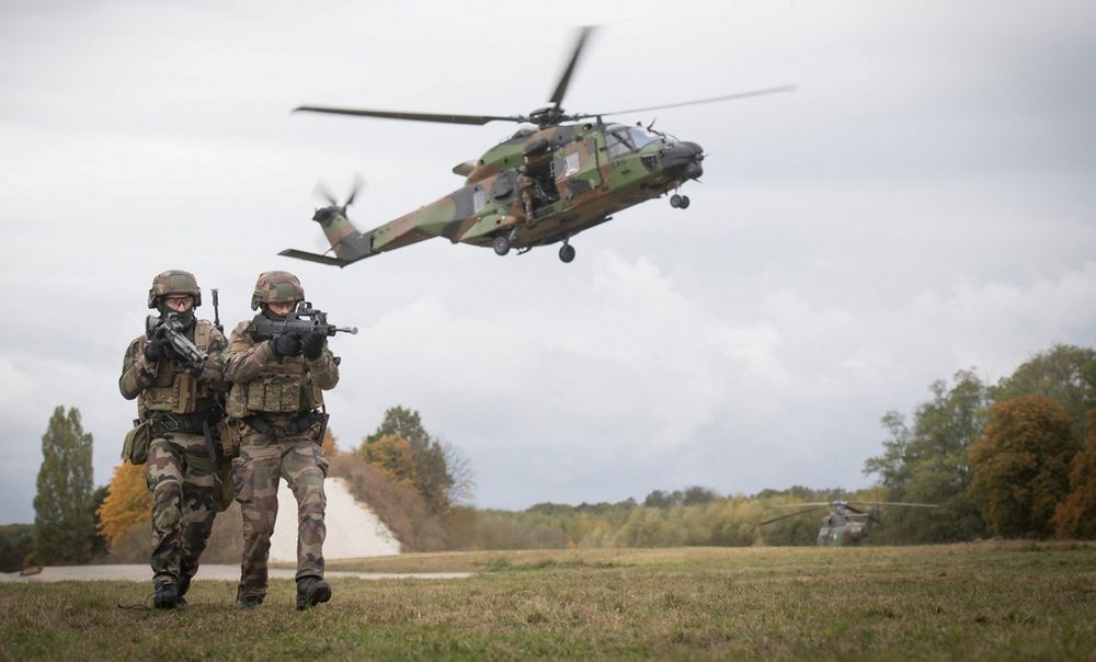 Two combat troops advance while an NH90 helicopter takes off in the background. 