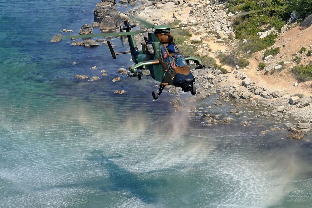 An Airbus Tiger HAD multi-role attack helicopter hovers over coastal terrain. 