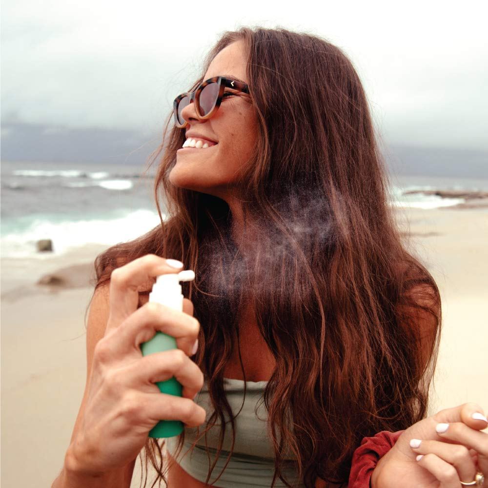 A woman applying the COOLA Organic Scalp & Hair Sunscreen Mist at the beach