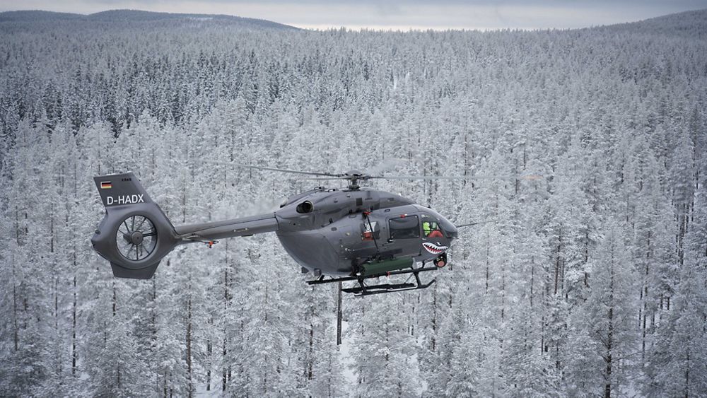 An Airbus H145 helicopter flies over snow-covered, wooded terrain. 