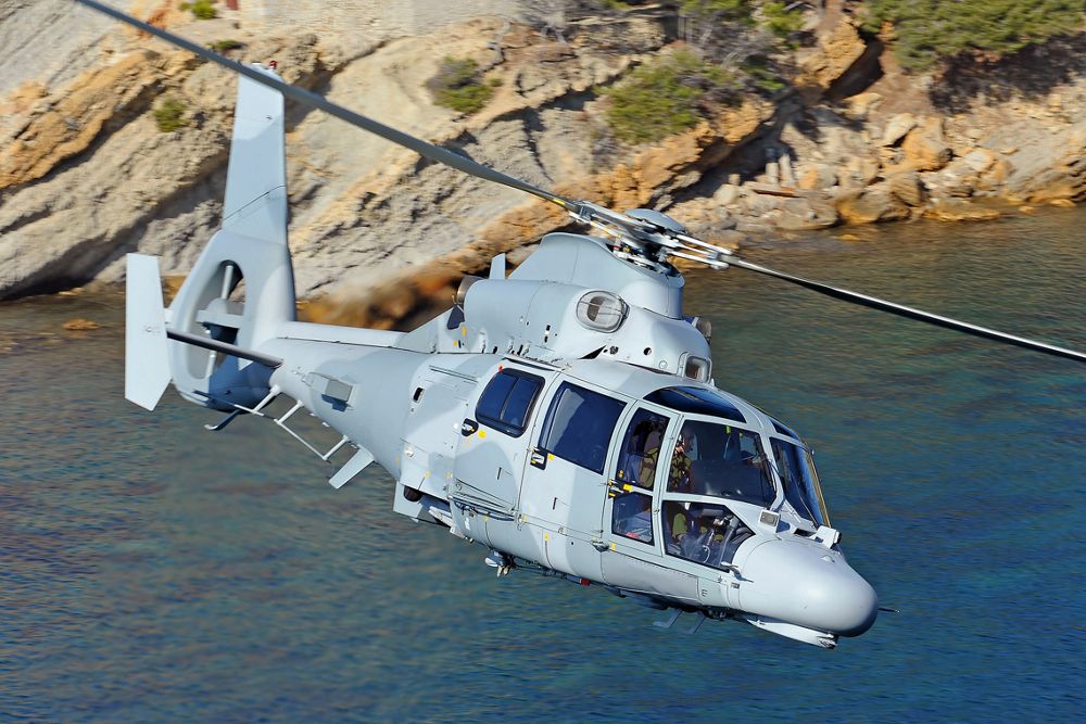 Side view of an Airbus AS565 MBe military helicopter flying over coastal terrain. 