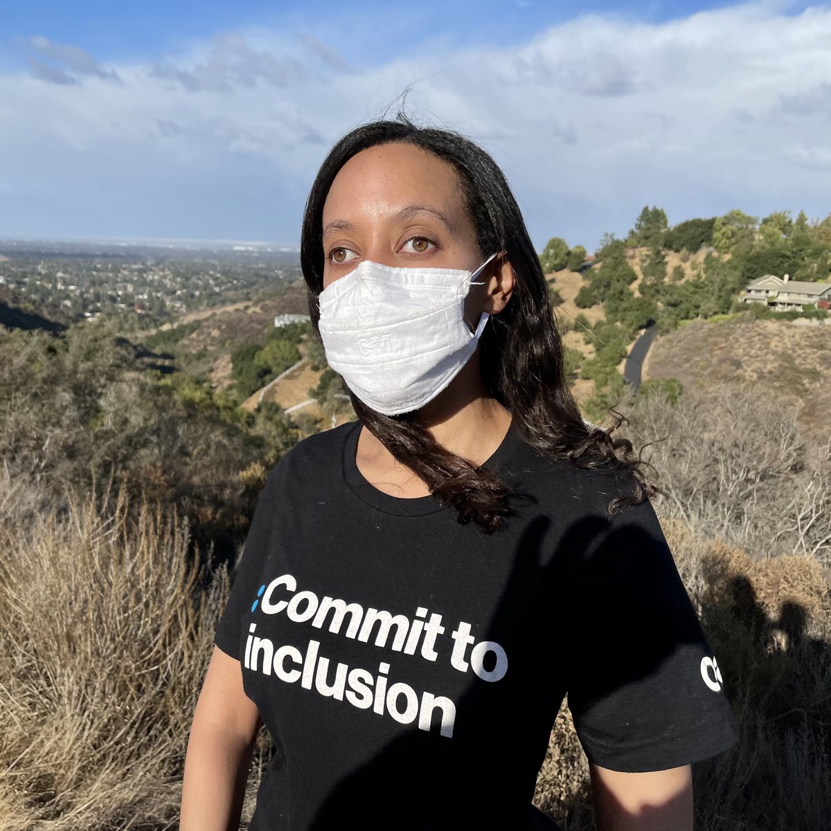 I’m wearing a black shirt with text that says, “Commit to Inclusion.” The white text goes with my white face mask. Behind me are yellow and green hills.