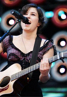 Higgins stands and plays an acoustic guitar with her left hand high on the fret board. She sings into a microphone. Her right arm and bottom of guitar are not in view. Background has large stage lights.