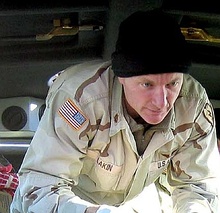 A caucasoid man in desert fatigues and a black knit cap is standing, bent-over towards the camera, and looking off center at an unpictured patient.