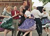 A group of dancers in colorful Western clothing promenading in a circle, with a man speaking into a microphone on a stage in the background.