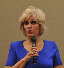 A photograph of a blond-haired, brown-eyed, middle-aged woman in a blue blouse holding a microphone.