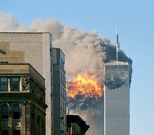 A monumental green copper statue of a woman with a torch stands on an island in front of a mainland where a massive plume of grey smoke billows among skyscrapers.