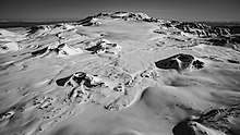 A snow-covered plateau with four small cone-shaped mounds in the foreground and a much larger flat-topped mountain in the background.