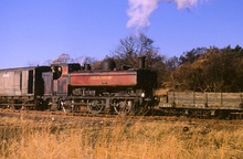 A pannier tank locomotive is pushing a shunter's wagon and pulling a guard's van. The locomotive is painted in a faded maroon with black and yellow piping, but much of the paint has peeled, revealing the black paint underneath. The lettering "LONDON TRANSPORT" is shown in yellow on the side of the pannier tank.