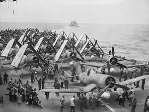 Black and white photo of a large number of single-engined monoplane aircraft on the deck of an aircraft carrier at sea. Another ship is visible in the background.