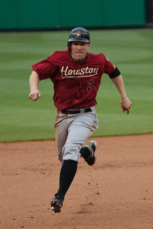 A white man in a red baseball uniform running bases. Looking into the camera, he has on a red baseball uniform reading "Houston" in yellow text with similarly-colored line under it. Below that to the runner's left hand side, there is a black "2" with a red outline. In addition to a black baseball with an orange star, he has on gray pants, black socks, and black cleats.