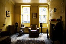 A wood panelled room, with polished wood floor, partly covered by a rug in the foreground. An armchair is between two sash windows, through which are the tops of spiked metal railings and, further away, Georgian brick houses with similar windows. On the mantlepiece above the fireplace are colourful spools of silks. In the foreground is a dummy, dressed in formal jacket, shirt and tie.