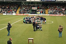 A view inside a football stadium. The winning team are posing together for a photograph, and there are photographers and journalists on the pitch.